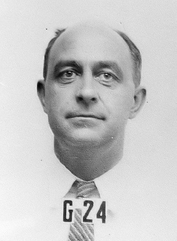 Enrico Fermi. Ein weißer Mann mit Glatze und Resthaar oberhalb der Schläfen. Er trägt ein weißes Hemd und eine Krawatte. Vor ihm befinden sich eine Kennnummer aus Plastikbuchstaben. "G 24".